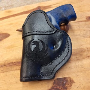 May include: Black leather holster with a stitched border and a Phoenix emblem. The holster holds a blue-handled handgun, resting on a wooden surface. The design is detailed and the leather appears to be high-quality.