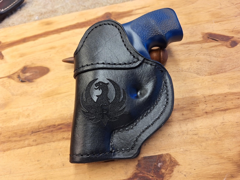 May include: Black leather holster with a stitched border and a Phoenix emblem. The holster holds a blue-handled handgun, resting on a wooden surface. The design is detailed and the leather appears to be high-quality.