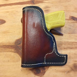 May include: A handcrafted leather holster with a two-tone brown and black finish. The holster features white stitching and a contoured design. A yellow object is partially visible inside the holster.