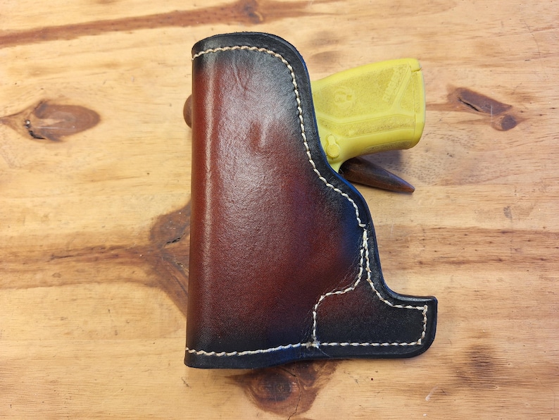May include: A handcrafted leather holster with a two-tone brown and black finish. The holster features white stitching and a contoured design. A yellow object is partially visible inside the holster.