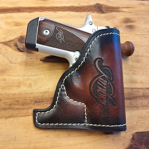 May include: A brown leather holster with white stitching, holding a silver and wood-grain handgun. The holster features a decorative embossed design and the name "Kimber".