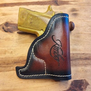 May include: A brown and black leather holster with white stitching, holding a gold-colored handgun. The holster has the name "Kimber" in a cursive font. The leather has a gradient effect, transitioning from black to brown.