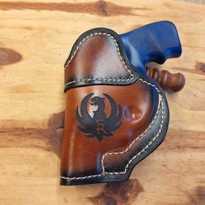 May include: A brown leather holster with a black stitched border and a black logo of a bird with the letters "SR" inside. The holster is designed to hold a blue handgun.
