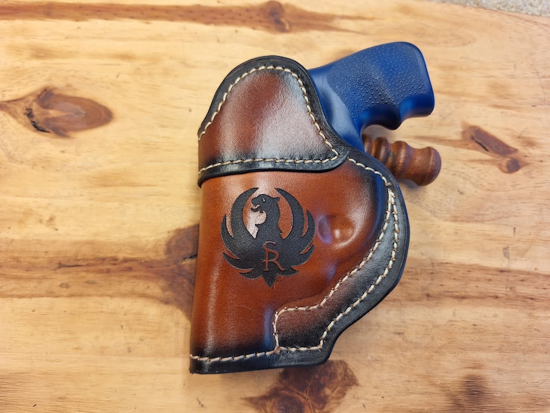 May include: A brown leather holster with a black stitched border and a black logo of a bird with the letters "SR" inside. The holster is designed to hold a blue handgun.