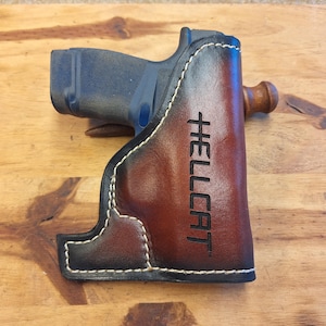 May include: A brown leather holster with white stitching and the word "HELLCAT" printed vertically. The holster is designed to hold a handgun, which is partially visible. The background is a wooden surface.