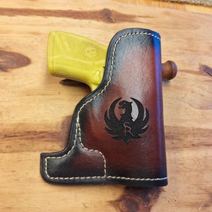 May include: A brown and black leather holster with white stitching, featuring a black emblem of a bird. A yellow handgun is partially visible inside the holster. The holster is resting on a wooden surface.
