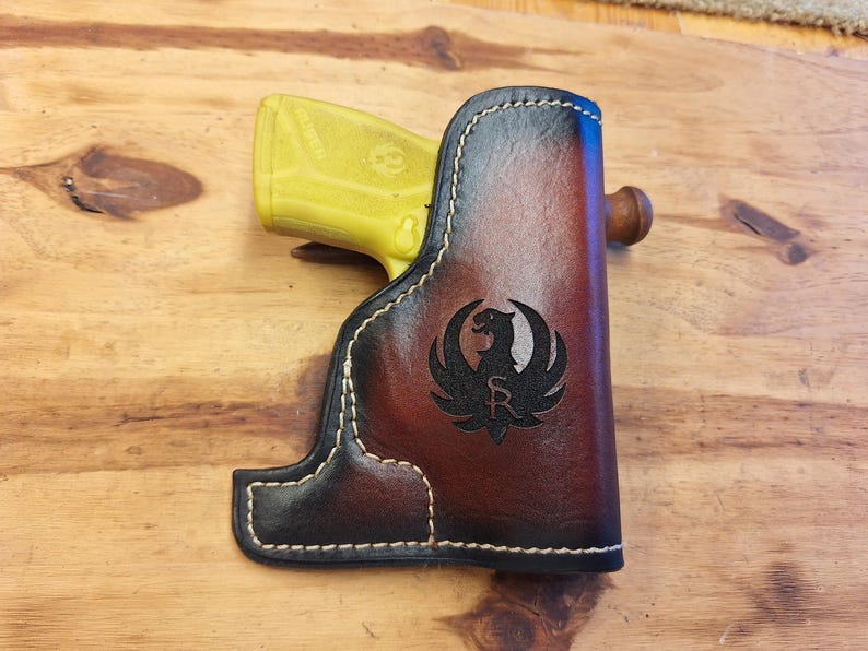 May include: A brown and black leather holster with white stitching, featuring a black emblem of a bird. A yellow handgun is partially visible inside the holster. The holster is resting on a wooden surface.