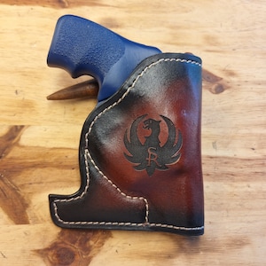 May include: A brown leather holster with a stitched edge and a black logo of a bird with the letters "SR" inside. The holster is designed to hold a blue handgun.