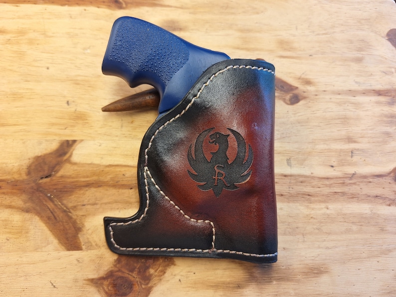 May include: A brown leather holster with a stitched edge and a black logo of a bird with the letters "SR" inside. The holster is designed to hold a blue handgun.