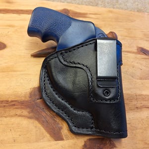 May include: A black leather holster with a silver clip holds a blue-handled handgun. The holster is stitched with a contrasting thread and sits on a wooden surface. The handgun is partially visible within the holster.