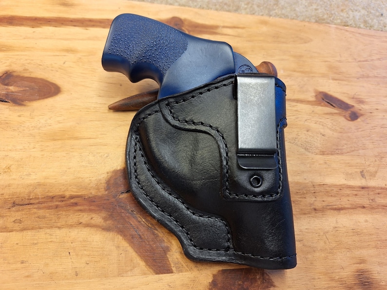 May include: A black leather holster with a silver clip holds a blue-handled handgun. The holster is stitched with a contrasting thread and sits on a wooden surface. The handgun is partially visible within the holster.