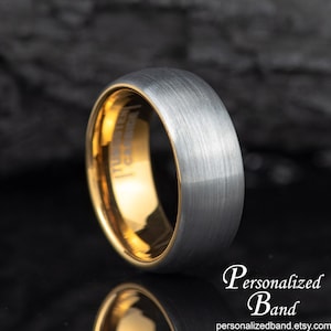 May include: A close-up of a tungsten carbide ring with a brushed silver exterior and a gold interior. The ring is on a reflective black surface. The text "Personalized Band" and the website address are in the image.