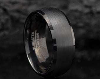 May include: A close-up of a black tungsten carbide ring. The ring has a brushed metal finish on the top surface and a polished, beveled edge. The words "Tungsten Carbide" are etched on the inside of the ring.