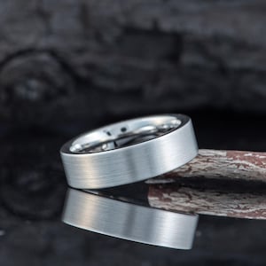 May include: A silver-colored ring with a brushed metal finish. The ring is set against a dark background, with its reflection visible below. The ring has a flat, wide band, and the interior appears to be a lighter shade of silver. The ring is a piece of jewelry.