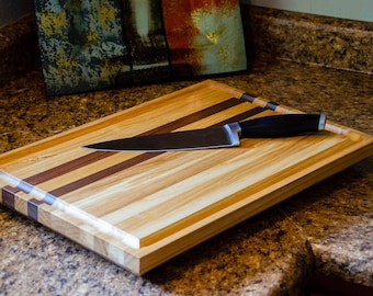 May include: A wooden cutting board with a natural wood finish and a black knife resting on top. The board has a multi-striped design with alternating light and dark wood.