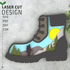 Könnte beinhalten: Ein Laser-Schnittdesign eines Wanderschuhs mit einer Bergszene im Inneren. Der Schuh ist braun und schwarz mit blauem Himmel, grünen Bäumen und einer gelben Sonne. Der Text "LASER CUT DESIGN" steht in grünen Buchstaben über dem Schuh.