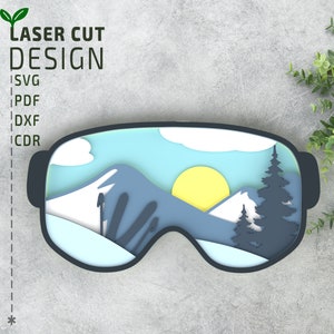 May include: A laser cut design of a pair of ski goggles with a mountain scene inside. The goggles are black with a blue lens. The mountain scene is made up of white snow, blue mountains, and a yellow sun.
