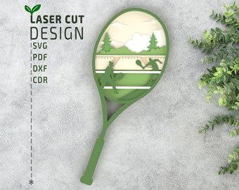 May include: A green laser cut design of a tennis racket with a scene of two people playing tennis inside the racket head. The scene includes a green tennis court, a white fence, and a green tree. The text "LASER CUT DESIGN" is above the racket.