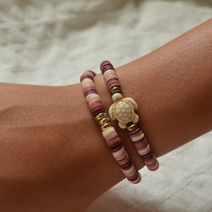 May include: Two bracelets made of pink, white, and gold beads with a white turtle charm. The bracelets are worn on a person's wrist.