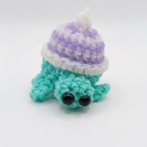 Può includere: Una tartaruga fatta a mano con elastici turchesi con un cappello viola e bianco. La tartaruga ha occhi di perline nere e un volant bianco intorno al cappello. Il giocattolo è fatto di piccoli elastici.