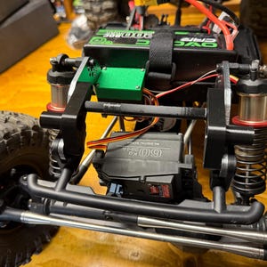 May include: Close-up of a black and green RC car chassis with large, textured tires. The chassis features a black frame, silver suspension components, and a green electronic component. The car has a black battery pack and a servo motor.