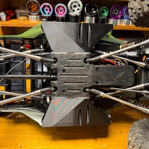 May include: Close-up of the underside of a black RC car chassis. The chassis features numerous silver suspension components, including rods and shocks. The car is on a wooden surface, with a shelf of colorful wheels in the background.