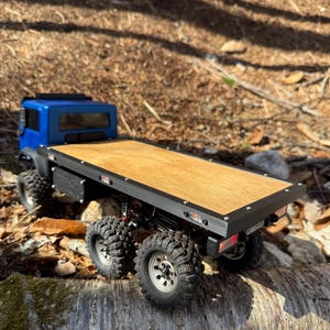 May include: A blue and black RC flatbed truck with a wooden bed. The truck has six large, knobby tires and is sitting on a log. The truck bed is made of wood and is surrounded by a black frame.