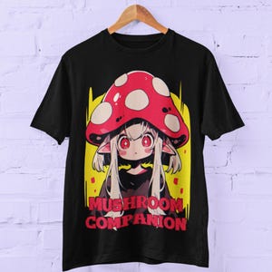 Può includere: T-shirt nera con un design a fumetto di un personaggio fungo. Il personaggio ha un cappello rosso e bianco a pois, grandi occhi rossi e capelli bianchi. Il testo "Mushroom Companion" è stampato in rosso e bianco.