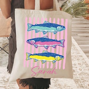 May include: Beige tote bag featuring three colorful fish illustrations on a pink striped background. The fish are blue, pink, and yellow. The name "Sarah" is written in pink script below the fish.