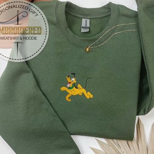 May include: An olive green sweatshirt with a crew neck and long sleeves. The sweatshirt features an embroidered image of Pluto, a yellow cartoon dog. A gold necklace with a pendant is visible. The words "EMBROIDERED SWEATSHIRT & HOODIE" are printed on a circular label.