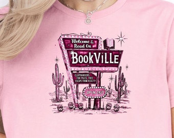 May include: Pink t-shirt featuring a retro-style sign that reads "Bookville." The sign includes the text "Welcome & Read On" and "Open 24/7." The design also includes cacti and stars.