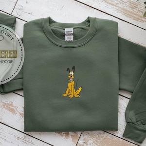 May include: An olive green sweatshirt with a cartoon Pluto embroidery. The design features a yellow dog with black ears and a black collar. The text "PERSONALIZED GIFT EMBROIDERED SWEATSHIRT & HOODIE" is in a circular design.