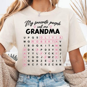 May include: Cream-colored t-shirt with the text "My favorite people call me GRANDMA" in black script. A word search grid includes names like Olivia and Roberta. The shirt is paired with denim shorts.