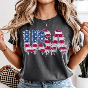 May include: Dark gray t-shirt with the word "USA" in a distressed, patriotic design. The letters are filled with the American flag's red, white, and blue colors. The shirt is short-sleeved and appears to be made of a soft material.