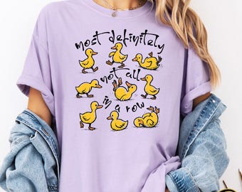 May include: A light purple t-shirt with the text "Most definitely not all in a row" and cartoon ducks. The ducks are yellow with orange beaks and feet. The shirt is a relaxed fit.