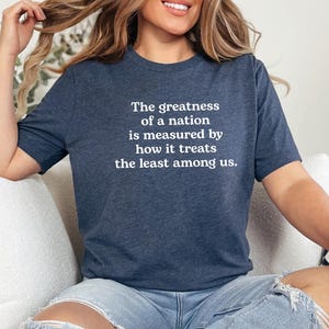 May include: Navy blue t-shirt with the quote "The greatness of a nation is measured by how it treats the least among us" in white text. The shirt is worn with ripped blue jeans. A gold bracelet is visible on the wrist.