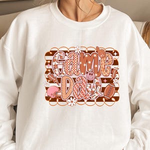 May include: White sweatshirt with a colorful "Game Day" graphic. The graphic features a football, stars, flowers, and a smiley face in shades of brown, pink, and orange. The sweatshirt is a casual, comfortable style.