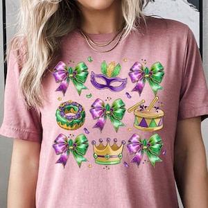 Puede incluir: Camiseta malva con un diseño colorido de Mardi Gras. El gráfico incluye lazos morados y verdes, una máscara, una corona, un tambor y un pastel decorado. El diseño es festivo y llamativo.