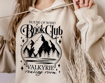 Peut inclure: Sweat à capuche beige avec un motif noir indiquant "HOUSE OF WIND Book Club VALKYRIE reading room" avec un design de montagne et de forêt. Le sweat à capuche a un cordon de serrage et une poche avant.
