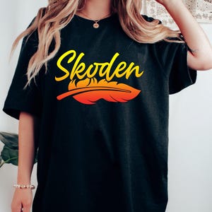 May include: A black oversized t-shirt with the text "Skoden" in yellow and orange lettering with a stylized orange and yellow feather underneath.