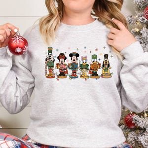May include: A light gray sweatshirt featuring a colorful graphic of Disney characters dressed as nutcrackers. The characters include Mickey Mouse, Minnie Mouse, Donald Duck, and Goofy. A red Christmas ornament is held in the hand.