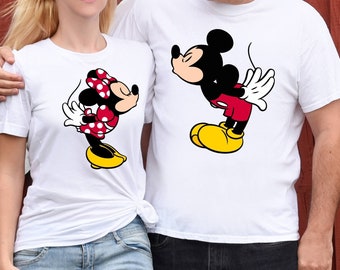 Könnte beinhalten: Ein Paar trägt weiße T-Shirts mit einem Cartoon-Bild von Mickey Mouse und Minnie Mouse. Der Mann trägt eine schwarze Jeans und die Frau eine blaue Jeans.