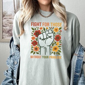 May include: A light green t-shirt with the text "FIGHT FOR THOSE WITHOUT YOUR PRIVILEGE" in orange and white. The design includes a raised fist surrounded by red and yellow flowers.