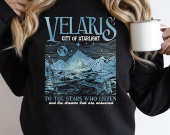 Peut inclure: Sweat-shirt noir avec un motif bleu et blanc. Le motif affiche "VELARIS CITY OF STARLIGHT" au-dessus d'un paysage montagneux, d'une lune et d'étoiles. Le texte en dessous dit "TO THE STARS WHO LISTEN and the dreams that are answered."