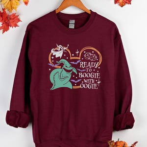 Pu&ograve; includere: Felpa bordeaux con una grafica di Oogie Boogie, pipistrelli e la scritta "Ready to Boogie with Oogie!". Il design &egrave; nei toni del verde, viola e arancione. La felpa &egrave; appesa a un gancio di legno.