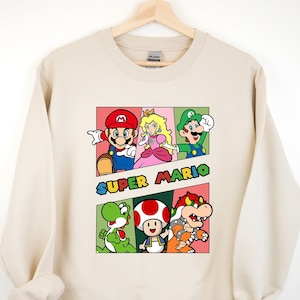 May include: A beige sweatshirt with a graphic print of characters from the Super Mario video game series. The characters include Mario, Luigi, Princess Peach, Yoshi, Toad, and Bowser. The text "SUPER MARIO" is printed in a colorful font.