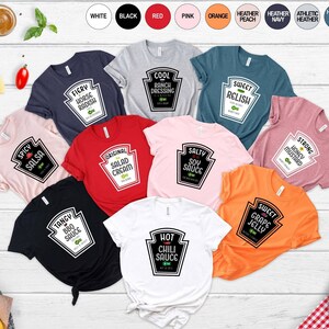 May include: Ten different t-shirts with various sauce-themed designs. Each shirt features a white ketchup bottle label with a different sauce name and description. The shirts are in various colors, including red, pink, orange, black, white, and heather grey.