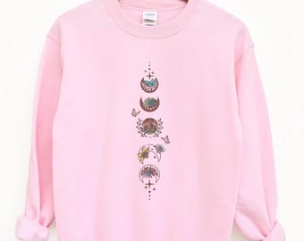 Peut inclure: Un sweat-shirt rose clair à col rond et manches longues. Il présente un motif vertical de phases lunaires avec des détails floraux et papillons. Le sweat-shirt est suspendu à un cintre en bois.
