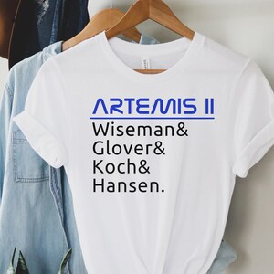 May include: White t-shirt with the text "ARTEMIS II" in blue, followed by the names Wiseman, Glover, Koch, and Hansen in black. The shirt is hanging on a wooden hanger with other clothing items in the background.