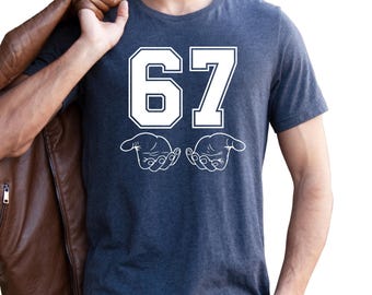 May include: Dark gray t-shirt with the number "67" in white block letters and two white hand illustrations. A brown leather jacket is draped over the shoulder. The person is wearing blue jeans.